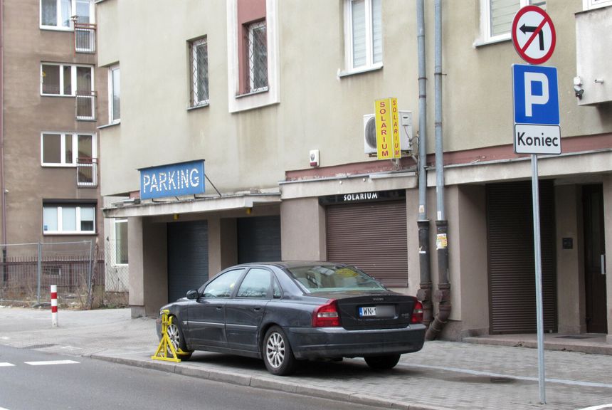 Zdjęcie przedstawia zaparkowany samochód na chodniku obok budynku z oznaczeniem „PARKING”.