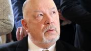 Mężczyzna w garniturze z szarymi włosami i wąsem, patrzy w górę, otoczony przez innych ludzi.