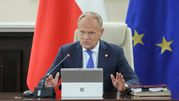 Mężczyzna w garniturze siedzi przy stole, mówi do mikrofonu, w tle flagi i dokumenty. Donald Tusk.