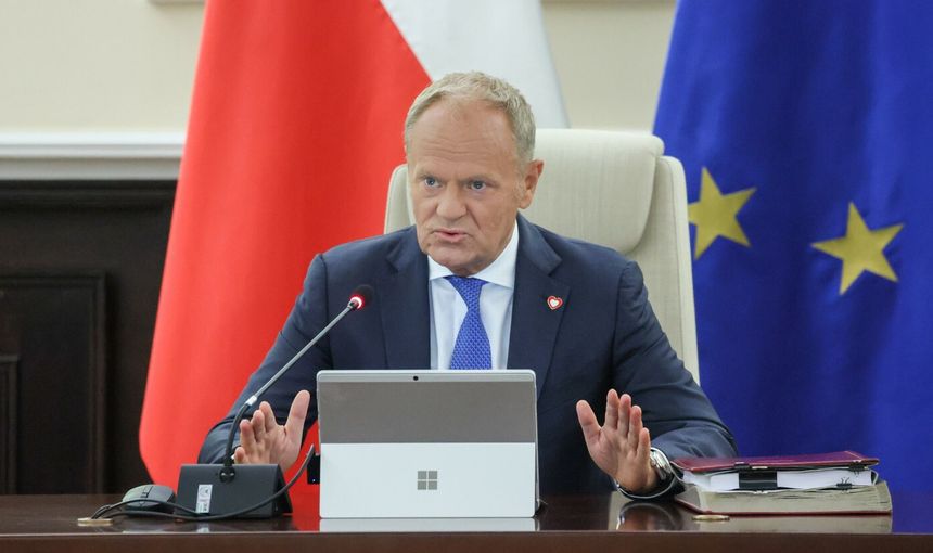 Mężczyzna w garniturze siedzi przy stole, mówi do mikrofonu, w tle flagi i dokumenty. Donald Tusk.