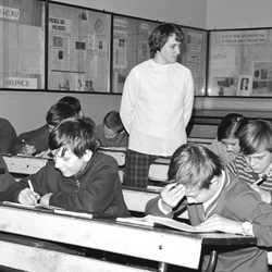 Zdjęcie czarno-białe z sali lekcyjnej w PRL-u. Uczniowie piszą, nauczycielka obserwuje z tyłu.