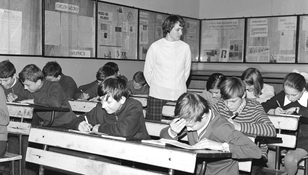 Zdjęcie czarno-białe z sali lekcyjnej w PRL-u. Uczniowie piszą, nauczycielka obserwuje z tyłu.