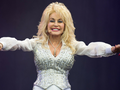 Na zdjęciu Dolly Parton w błyszczącym kostiumie, entuzjastycznie machająca rękami na scenie.