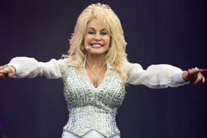 Na zdjęciu Dolly Parton w błyszczącym kostiumie, entuzjastycznie machająca rękami na scenie.