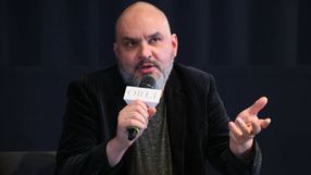 Mężczyzna w ciemnym garniturze, trzyma mikrofon, gestykulując podczas dyskusji.
