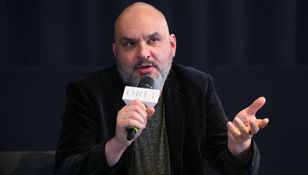 Mężczyzna w ciemnym garniturze, trzyma mikrofon, gestykulując podczas dyskusji.