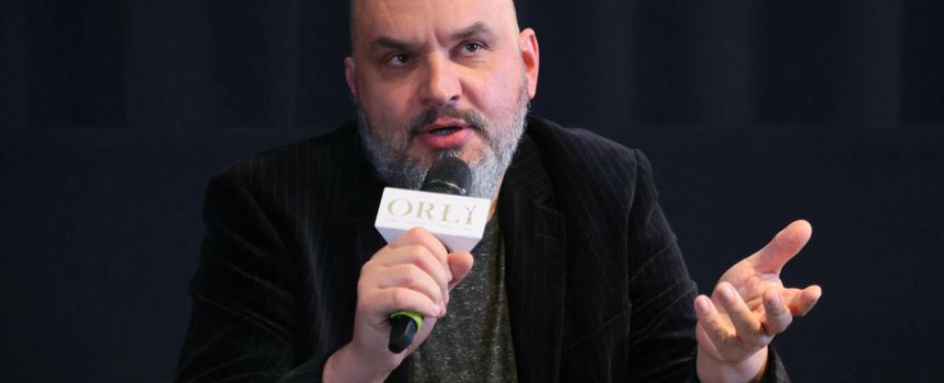Mężczyzna w ciemnym garniturze, trzyma mikrofon, gestykulując podczas dyskusji.