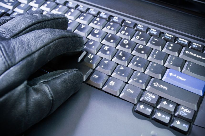 Na klawiaturze laptopa widać dłoń w czarnej rękawicy. W tle ciemny laptop.