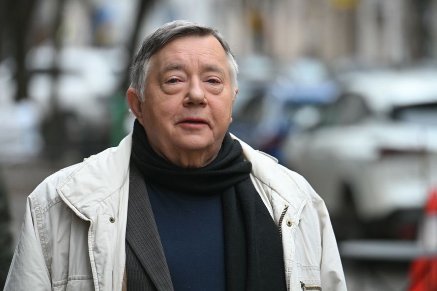Mężczyzna w jasnej kurtce i czarnym szalu spaceruje ulicą w mieście.