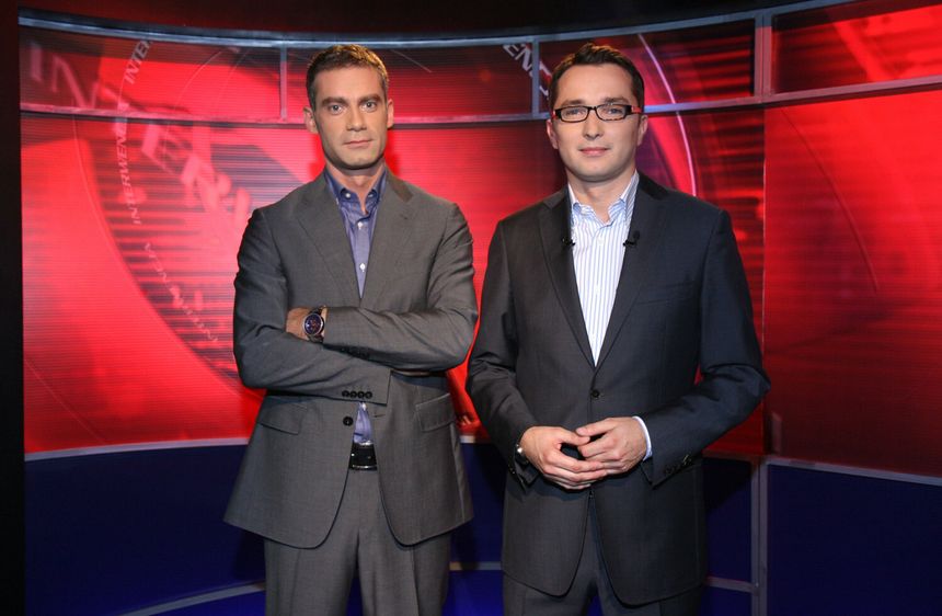 Dwaj mężczyźni w garniturach stoją przed czerwonym tłem w studiu telewizyjnym.