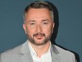 Mężczyzna w jasnym garniturze i białej koszuli, uśmiechający się na tle ciemnej ściany.