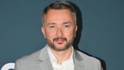 Mężczyzna w jasnym garniturze i białej koszuli, uśmiechający się na tle ciemnej ściany.