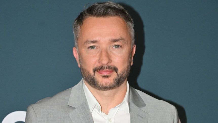 Mężczyzna w jasnym garniturze i białej koszuli, uśmiechający się na tle ciemnej ściany.