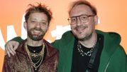 Na zdjęciu dwóch mężczyzn uśmiecha się, stojąc blisko siebie na tle pomarańczowej ściany.