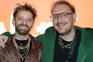 Na zdjęciu dwóch mężczyzn uśmiecha się, stojąc blisko siebie na tle pomarańczowej ściany.