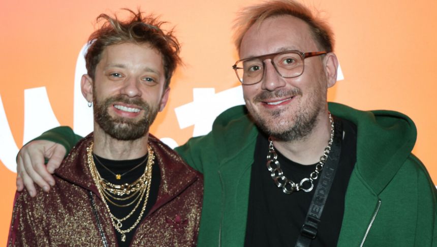 Na zdjęciu dwóch mężczyzn uśmiecha się, stojąc blisko siebie na tle pomarańczowej ściany.