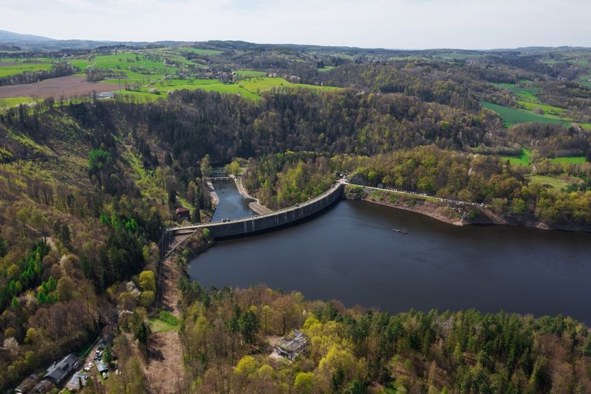 Widok na zaporę w zielonym, górzystym terenie, otoczoną wodą i lasami.