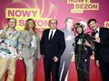 Grupa pięciu osób w kolorowych strojach pozująca na tle różowego tła z napisem 