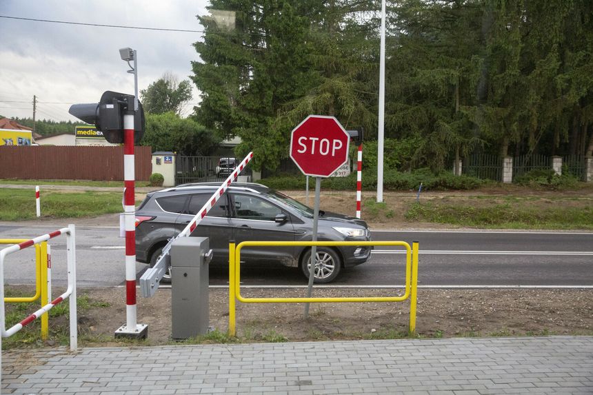 Samochód przejeżdża przez przejazd kolejowy obok znaku „STOP” i szlabanów przy drodze. W tle drzewa.