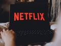 Na zdjęciu laptop z wyświetlonym logo Netflix na czarnym tle, ręka w zasięgu ekranu.