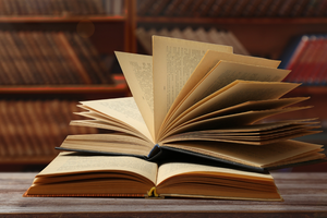 Otwarte książki na drewnianym stole, w tle rozmyta biblioteka z regałami.