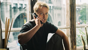 Na zdjęciu mężczyzna rozmawia przez telefon, siedząc przy dużym oknie. W tle widać miasto.