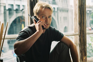 Na zdjęciu mężczyzna rozmawia przez telefon, siedząc przy dużym oknie. W tle widać miasto.