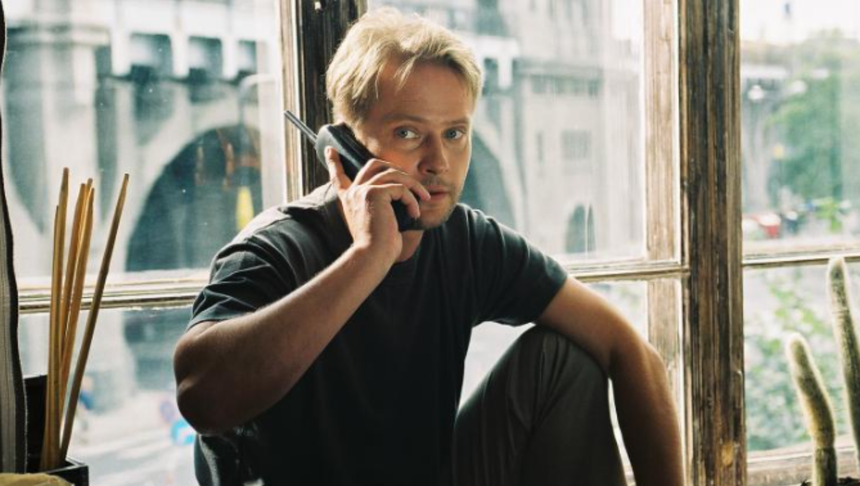 Na zdjęciu mężczyzna rozmawia przez telefon, siedząc przy dużym oknie. W tle widać miasto.