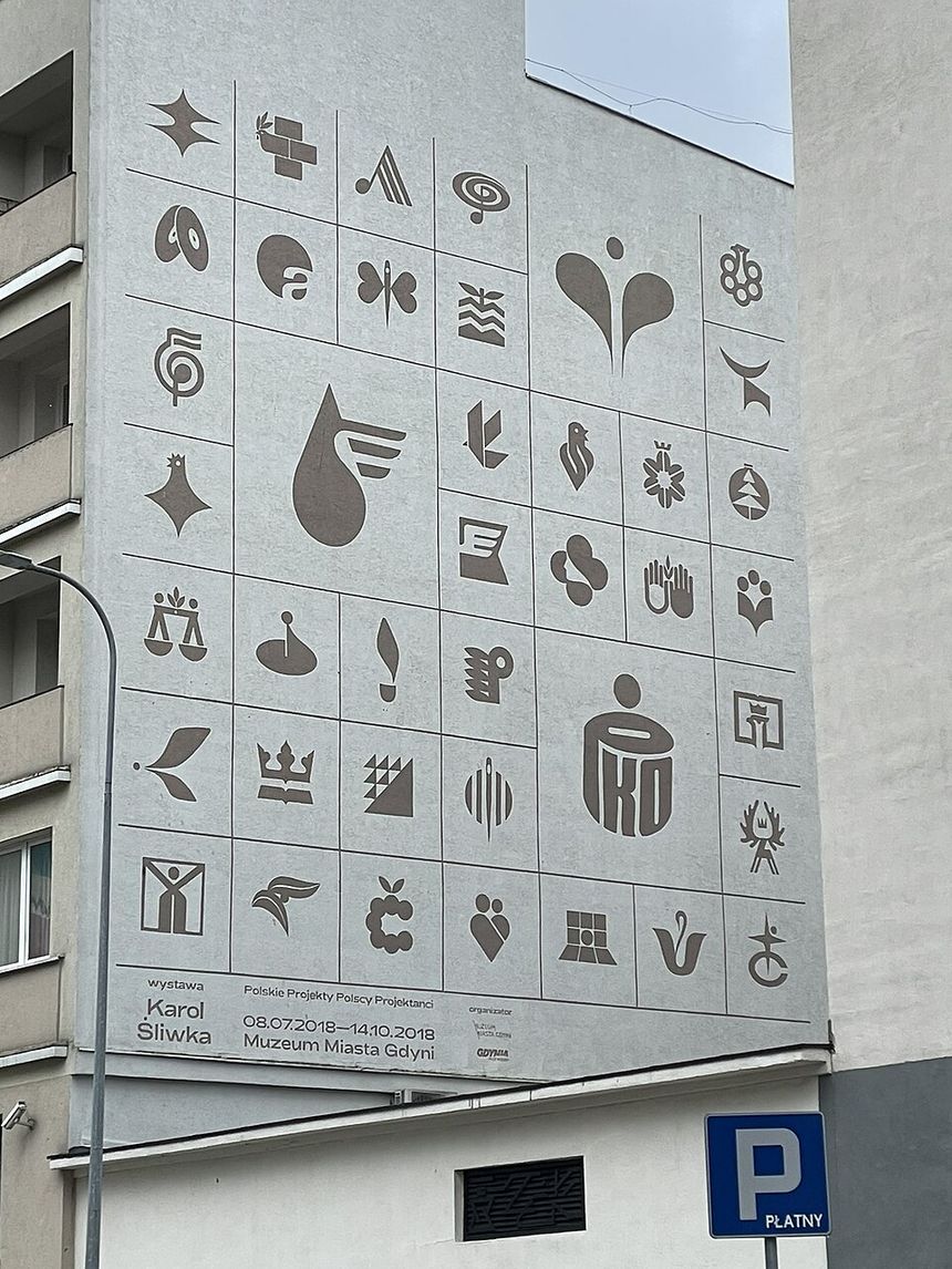 Na ścianie budynku widoczne różnorodne symbole oraz napisy związane z wystawą.