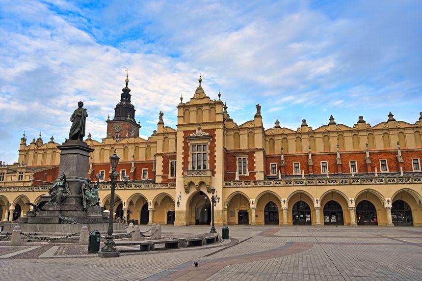 Widok na Sukiennice w Krakowie z pomnikiem w centrum oraz niebem w tle.