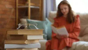 Na pierwszym planie stos książek z okularami, w tle kobieta czytająca książkę.