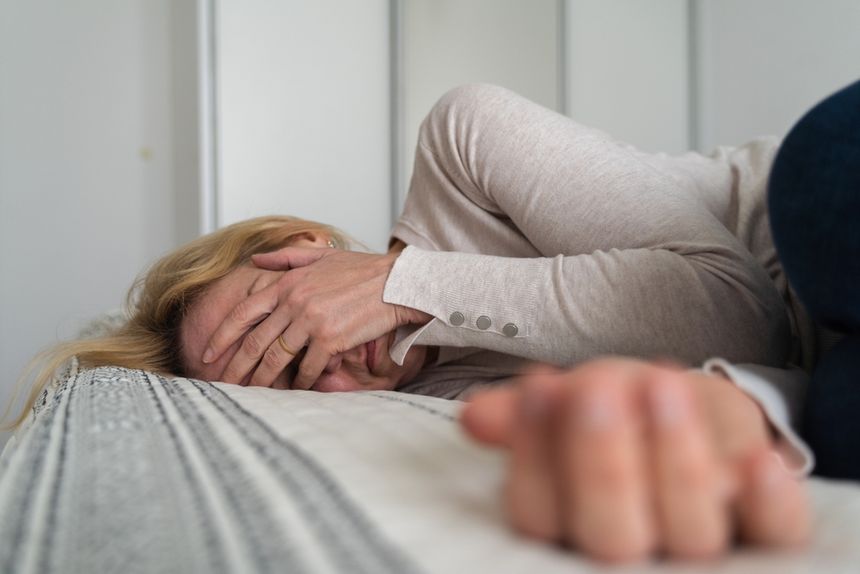 Kobieta w jasnym swetrze leży na łóżku, zakrywając twarz dłońmi w wyrazie smutku.
