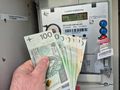 Zdjęcie przedstawia rękę trzymającą banknoty przy liczniku energii elektrycznej.