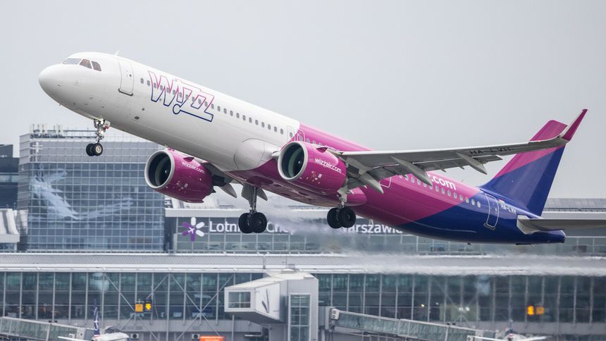Samolot linii Wizz Air startujący z lotniska, w tle budynki terminalu. Szare niebo.