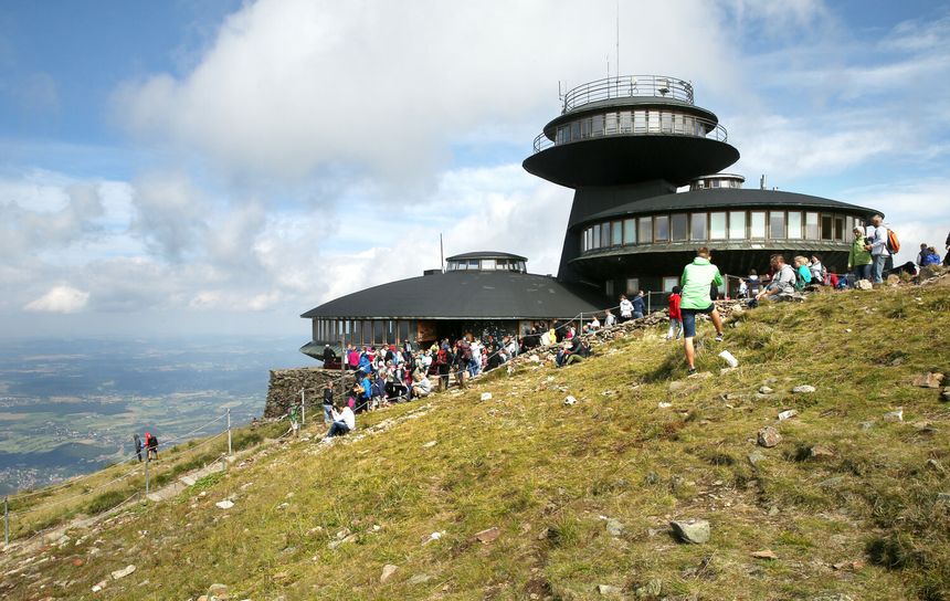 Widok na budynek górski z grupą ludzi na zielonej strefie i chmurami na niebie.