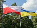 Flagi Polski i Ukrainy powiewają obok siebie na tle zielonych drzew i nieba.
