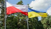 Flagi Polski i Ukrainy powiewają obok siebie na tle zielonych drzew i nieba.