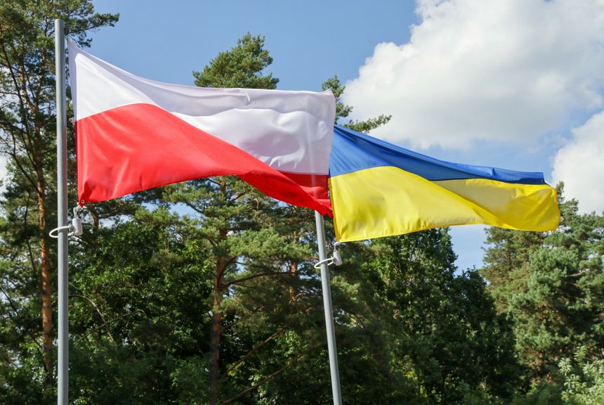 Flagi Polski i Ukrainy powiewają obok siebie na tle zielonych drzew i nieba.