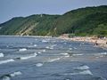 Plaża nadmorska, falujące morze, ludzie w wakacyjnych ubiorach, zieleń w tle.