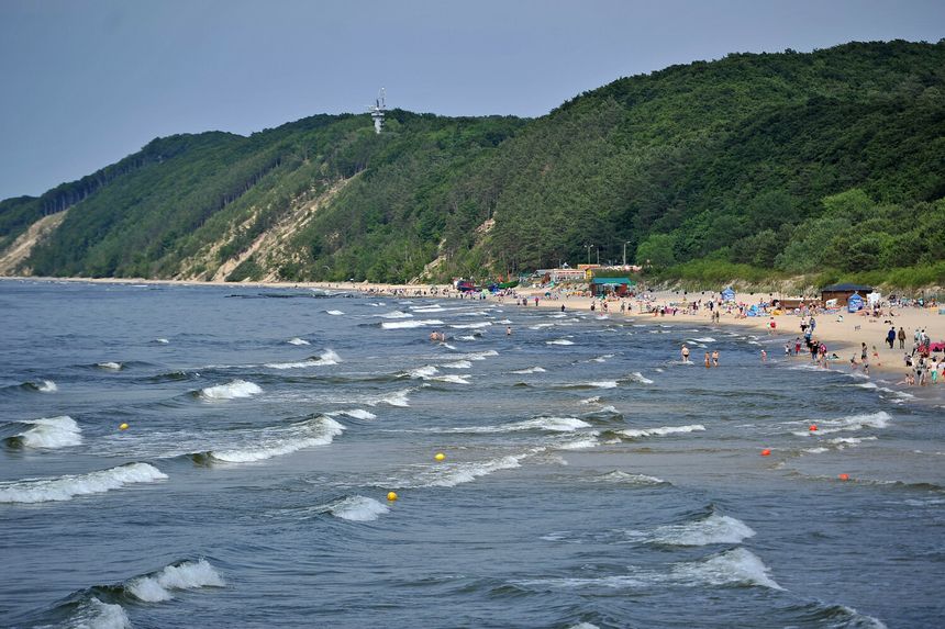Plaża nadmorska, falujące morze, ludzie w wakacyjnych ubiorach, zieleń w tle.