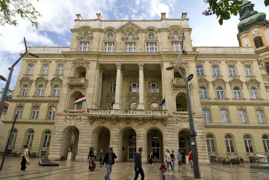 Historyczny budynek z kolumnami, nad nim niebo z chmurami, uliczni przechodnie.