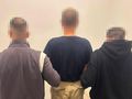Grupa trzech mężczyzn odwrócona tyłem, w tle jednolita ściana. Na jednym z nich widoczny napis 