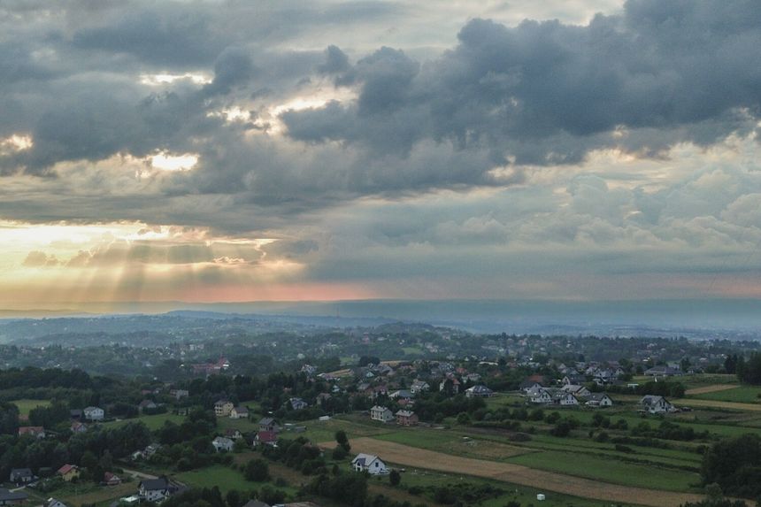 Krajobraz z zielonymi wzgórzami, domami i ciemnymi chmurami na tle zachodzącego słońca.