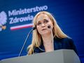 Kobieta z blond włosami przemawia na konferencji prasowej, tło z logo Ministerstwa Zdrowia.