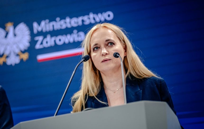 Kobieta z blond włosami przemawia na konferencji prasowej, tło z logo Ministerstwa Zdrowia.