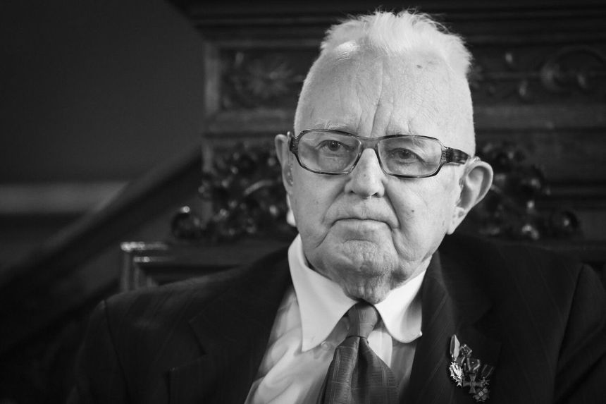 Mężczyzna w okularach, w eleganckim garniturze, siedzi w stylowym wnętrzu, poważny wyraz twarzy.