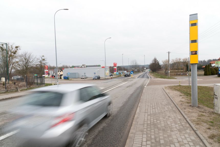 Widok ulicy z ruchem samochodowym, widoczny fotoradar, szare niebo. Brak osób.