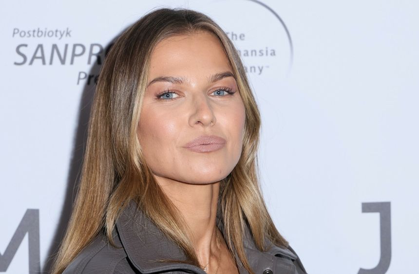 Anna Lewandowska z długimi włosami w jasnej kurtce, stoi na tle białego tła z logiem wydarzenia.