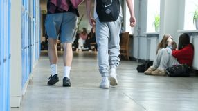 Dwa młode osoby w dresach idą korytarzem, w tle grupy uczniów siedzących w szkole.