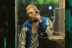 Mężczyzna w telefonie automatycznym, w dresie, z brodą i krótkimi włosami, skupiony na rozmowie.